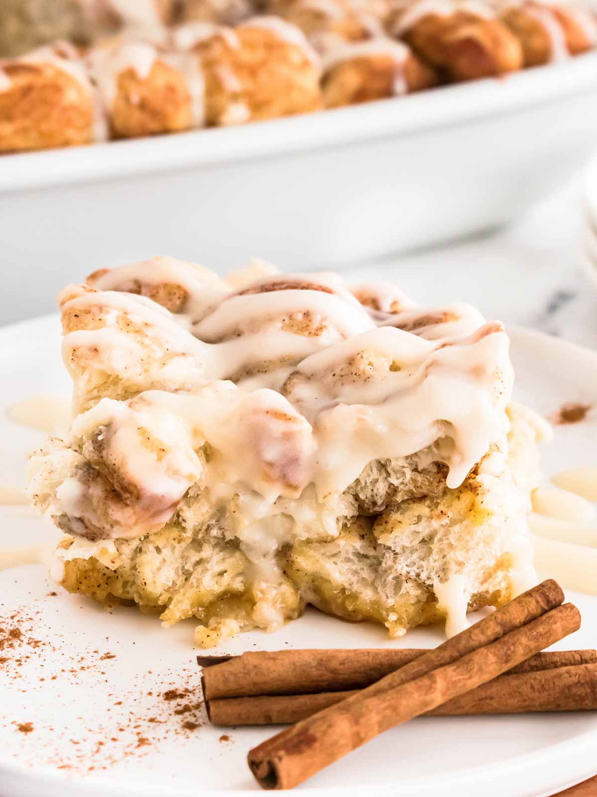 frontal view of cinnamon roll casserole slice on white plate with cinnamons sticks in front