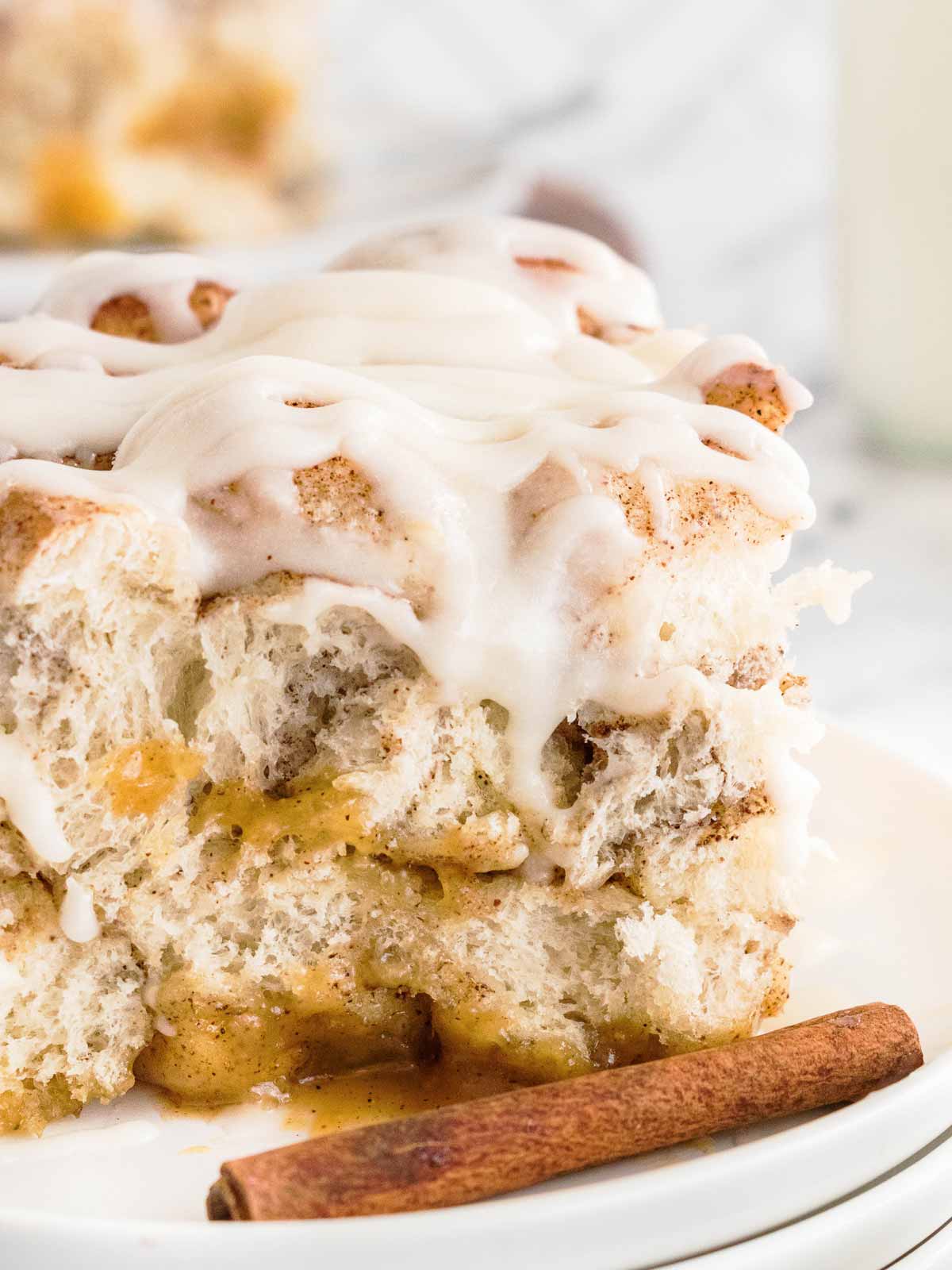 frontal close up view of a slice of cinnamon roll casserole on a white plate with cinnamon sticks