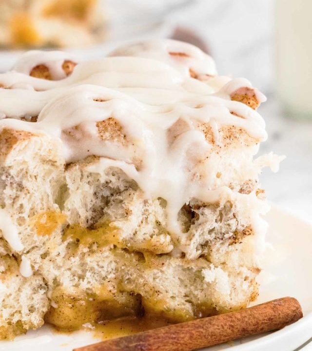 frontal close up view of a slice of cinnamon roll casserole on a white plate with cinnamon sticks